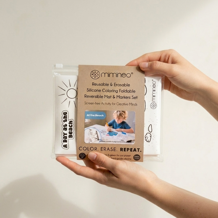 A person holds a packaged Mimneo reusable silicone coloring foldable set against a plain light background. The product is in a clear plastic sleeve with a tan paper label and white sleeves printed with black graphics and text.