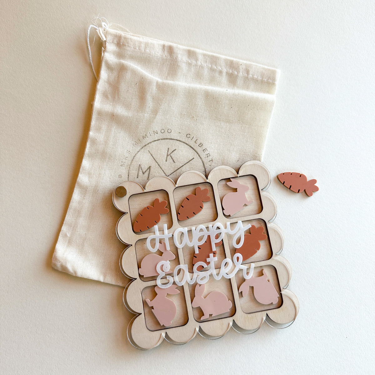 MKS Miminoo Easter Tic Tac Toe game with wooden board, white 'Happy Easter' overlay, pink bunny and brown carrot game pieces, and a branded canvas storage bag, all on a light background.