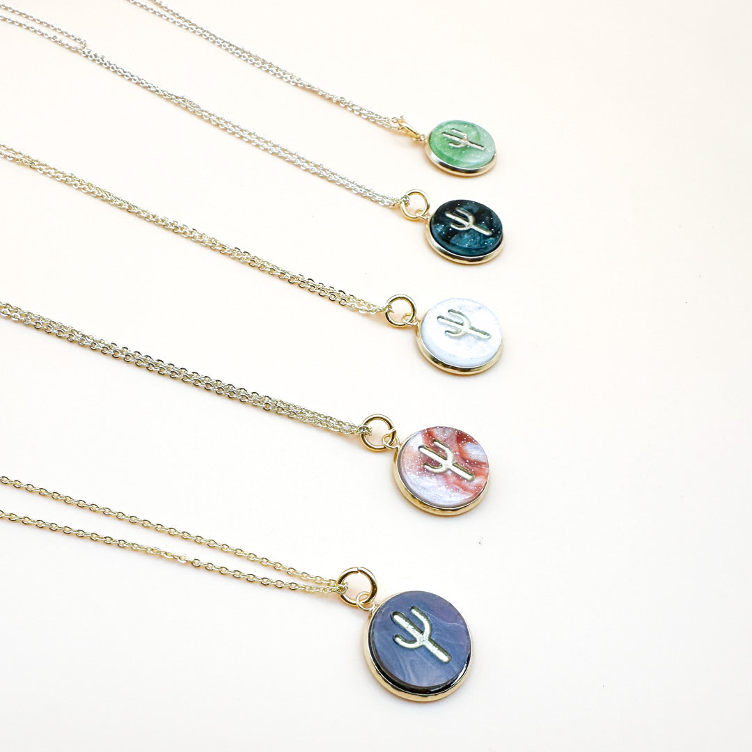A row of five pearl acrylic gold cactus necklaces, each with a different colored marbled acrylic pendant (green, dark blue, white, red, and purple), showcasing a gold cactus silhouette on a delicate gold chain by Miminoo