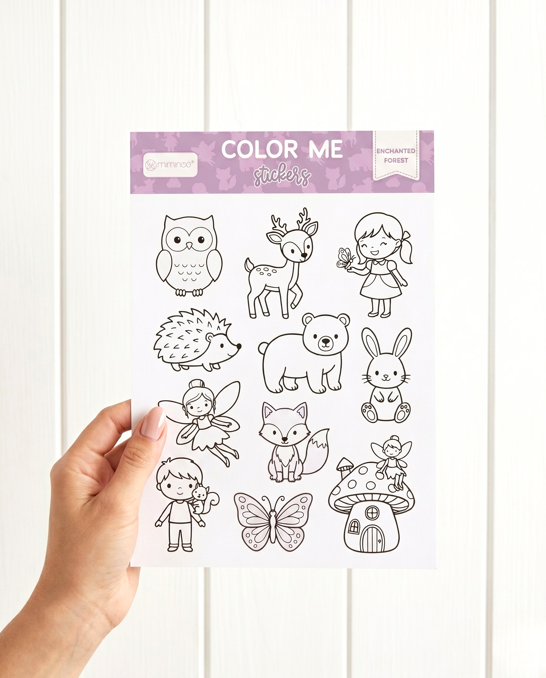 A hand holding a MKS Miminoo Color Me Enchanted Forest sticker sheet, featuring outline illustrations of an owl, deer, girl with butterfly, hedgehog, bear, bunny, fairy, fox, boy with squirrel, butterfly, and mushroom house, all ready for coloring.