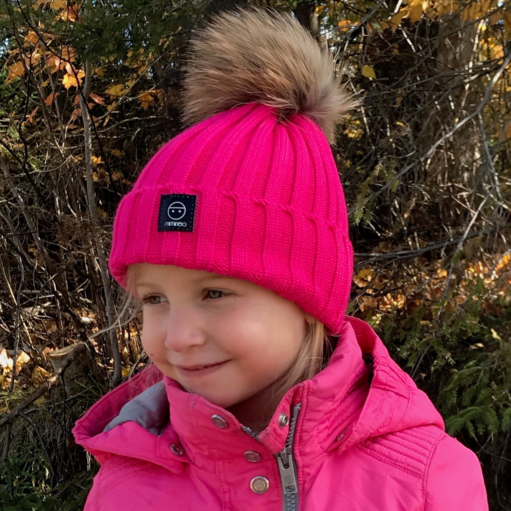 A young girl wearing a bright pink ribbed knit beanie with a brown faux-fur pom-pom and a matching pink winter jacket, standing outdoors in front of trees with autumn foliage.