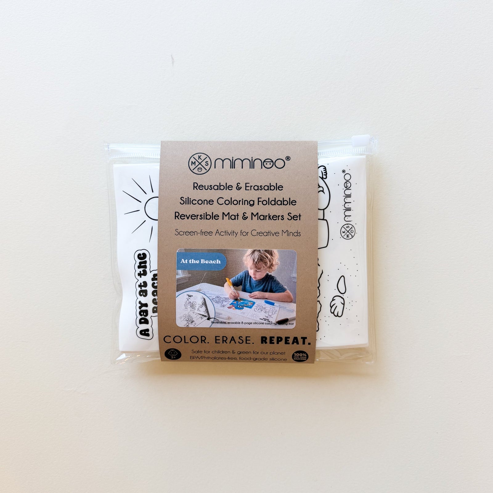 A packaged Minineeo Reversible Mat & Markers Set on a plain off-white background. The set includes a white silicone coloring mat with black line drawings and a white marker pad with black text, wrapped in clear plastic with a brown paper label.